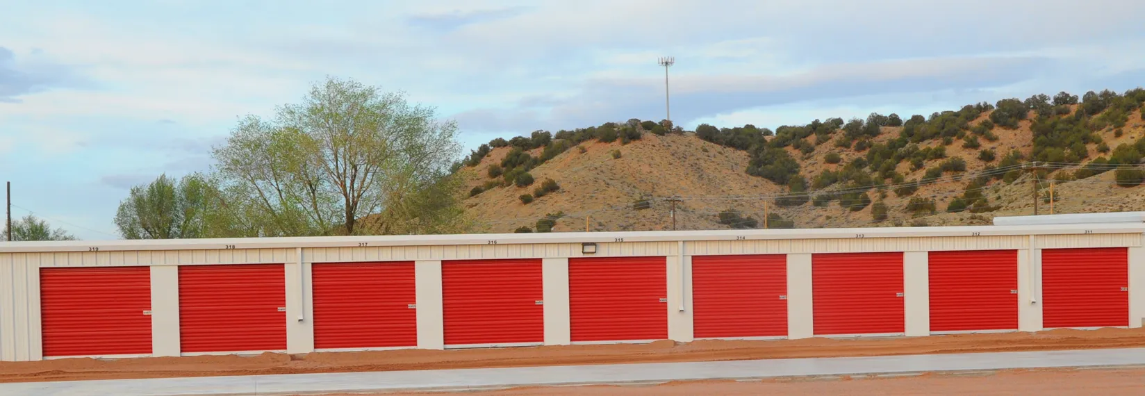 A long row of bright red roll-up storage doors on white steel buildings at Cañon City Self Storage, with the Colorado foothills rising behind them.