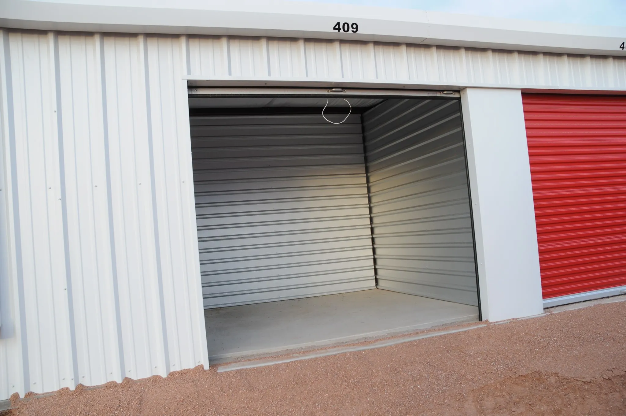 An open, empty storage unit with a swept concrete floor, clean corrugated steel walls and a red roll-up door on the neighbouring unit.
